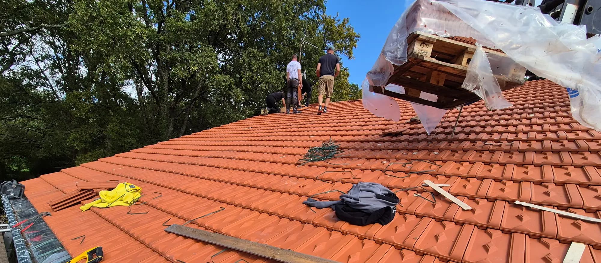 Trois couvreurs au travail sur une toiture en tuiles, une palette de tuiles hélitreuillée au-dessus du faîtage