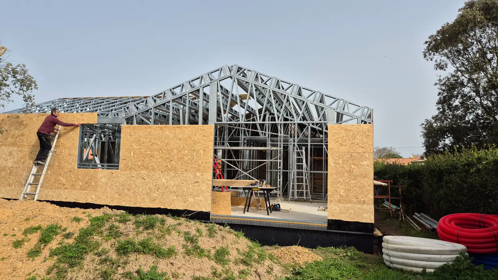 Une maison à ossature métallique en construction, un compagnon pose les panneaux de façade depuis une échelle